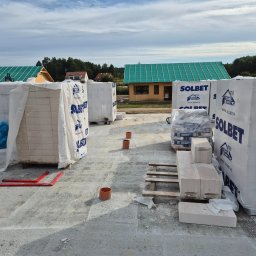 Jaroslaw Rudzki - Budowa domu w trakcie: bloczki Solbet na paletach, widoczny szkielet budynku z zielonym dachem, rury kanalizacyjne. Fundamenty w surowym stanie.