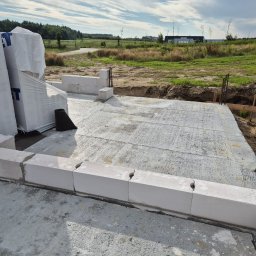 Jaroslaw Rudzki - Wylana płyta fundamentowa z częściowo ułożonymi bloczkami Ytong na obrzeżach, w tle widok na łąkę i budynek. Etap budowy domu.