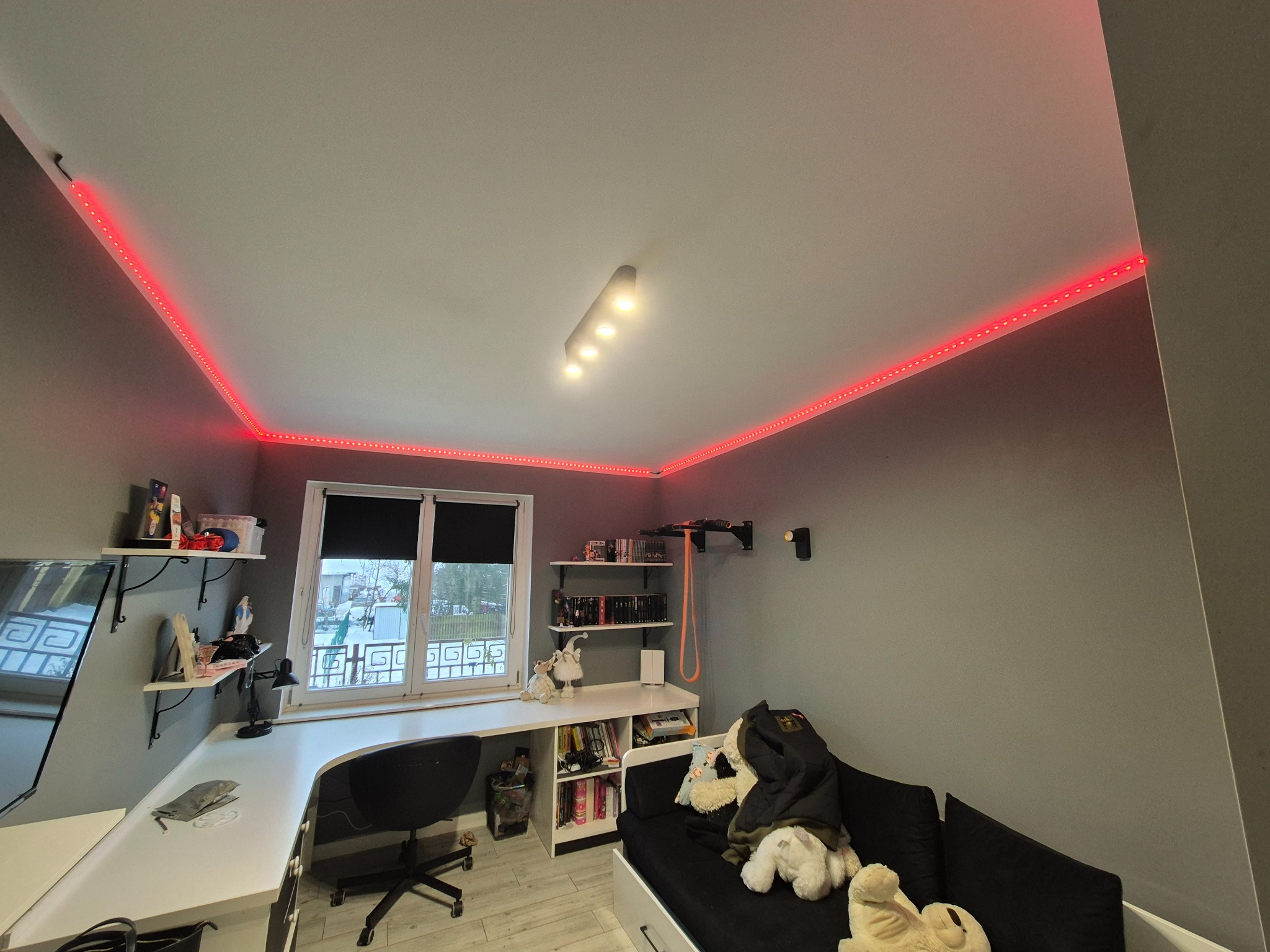 Pokój młodzieżowy z szarą ścianą, oświetleniem LED na suficie i biurkiem pod oknem. Widoczne półki, kanapa i zabawki. Nowoczesny design.