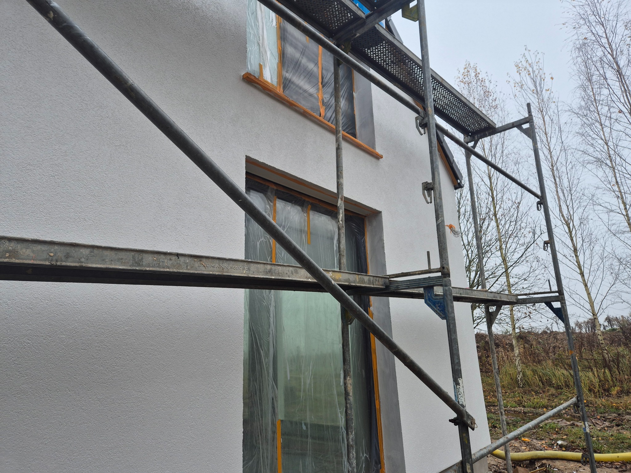 Elewacja domu w trakcie prac wykończeniowych, zabezpieczone okna folią, widoczne rusztowania. Świeżo nałożony tynk o fakturze baranka.