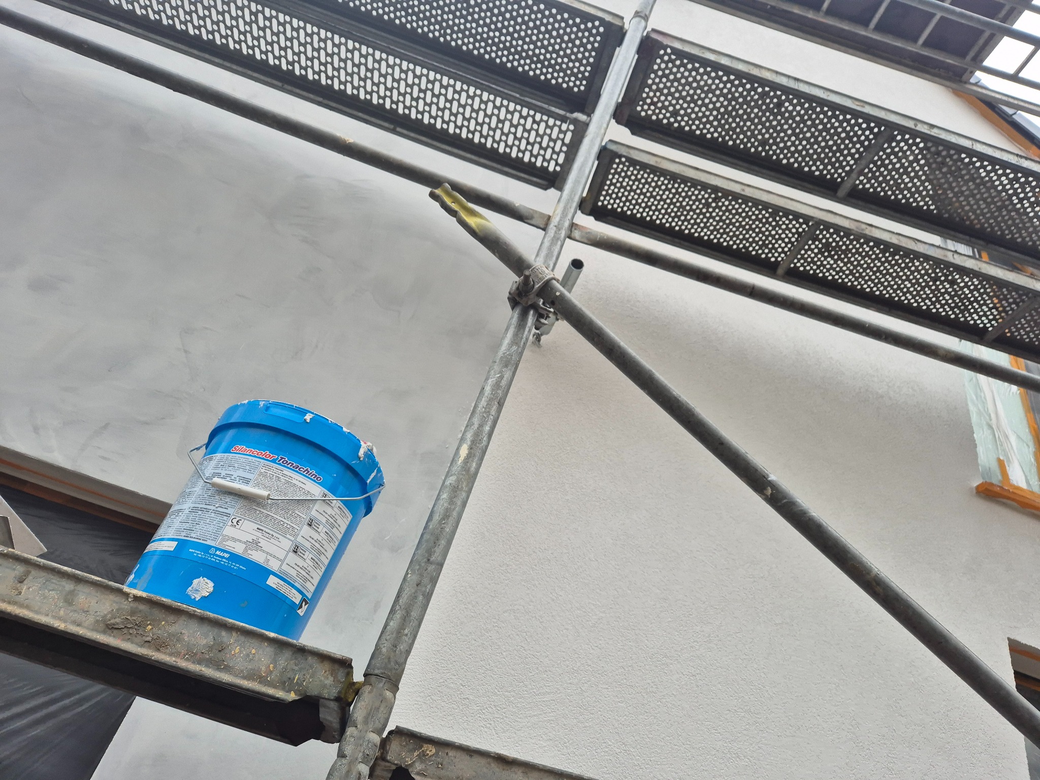 Ściana budynku po nałożeniu tynku strukturalnego, widoczna z rusztowania. Na rusztowaniu stoi wiadro z masą tynkarską. Ujęcie z dołu, podkreślające fakturę ściany.