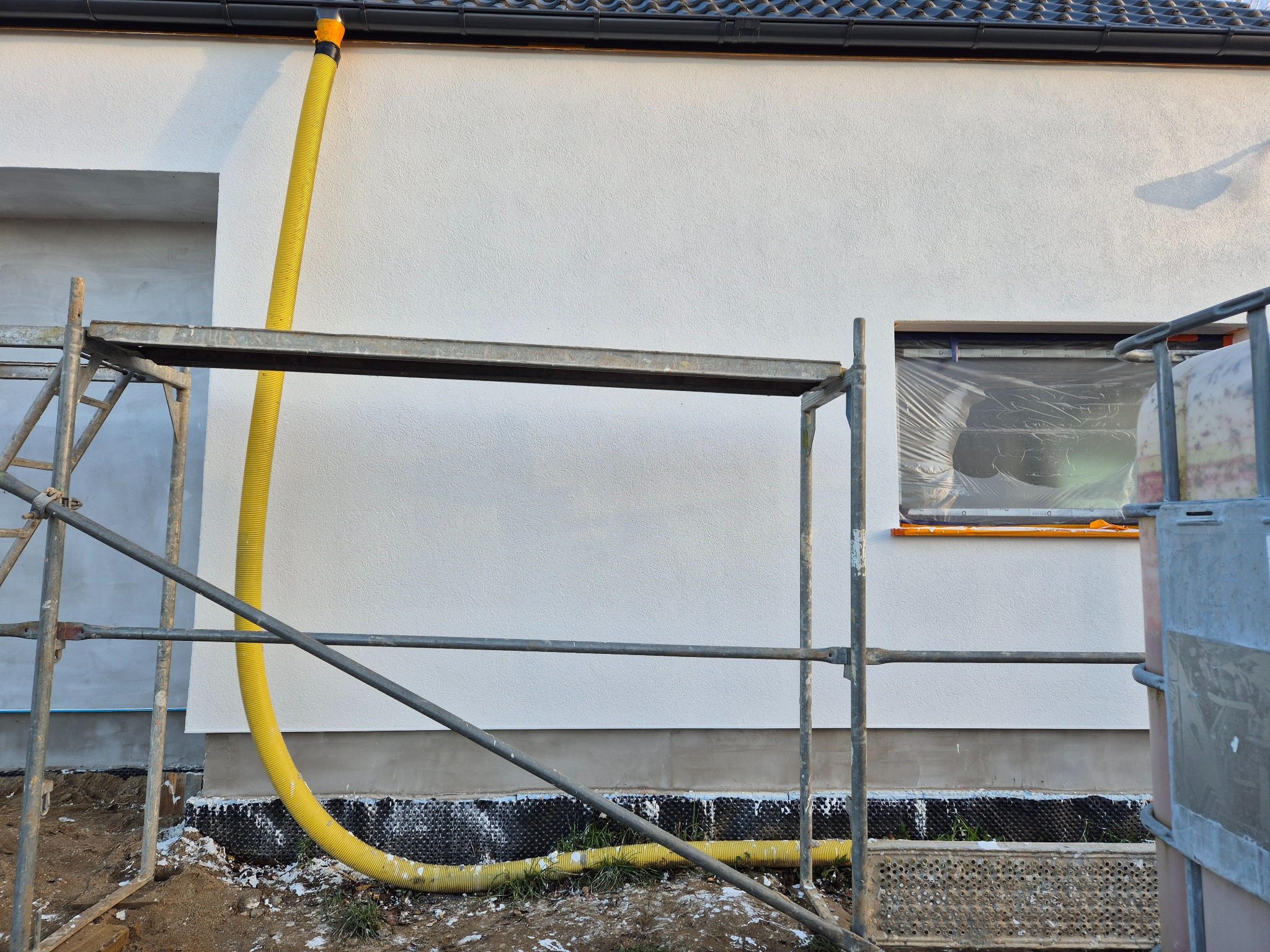 Elewacja domu w Lidzbarku Warmińskim w trakcie prac. Widoczne rusztowanie, żółty wąż wentylacyjny i okno zabezpieczone folią. Świeżo otynkowana ściana.