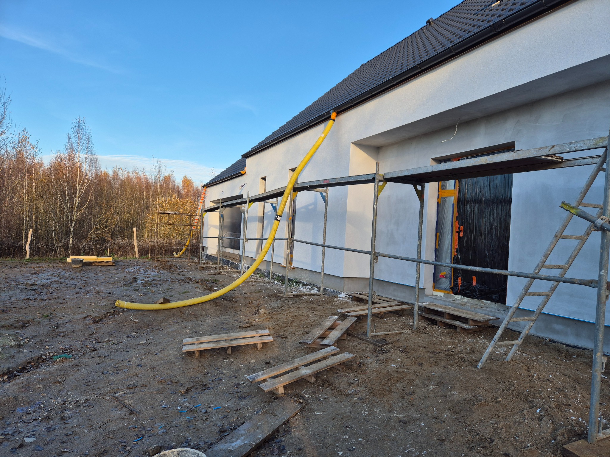 Elewacja domu w trakcie prac wykończeniowych, widoczne rusztowania, żółty wąż wentylacyjny i fragment lasu w tle. Świeżo otynkowana ściana w jasnym odcieniu.