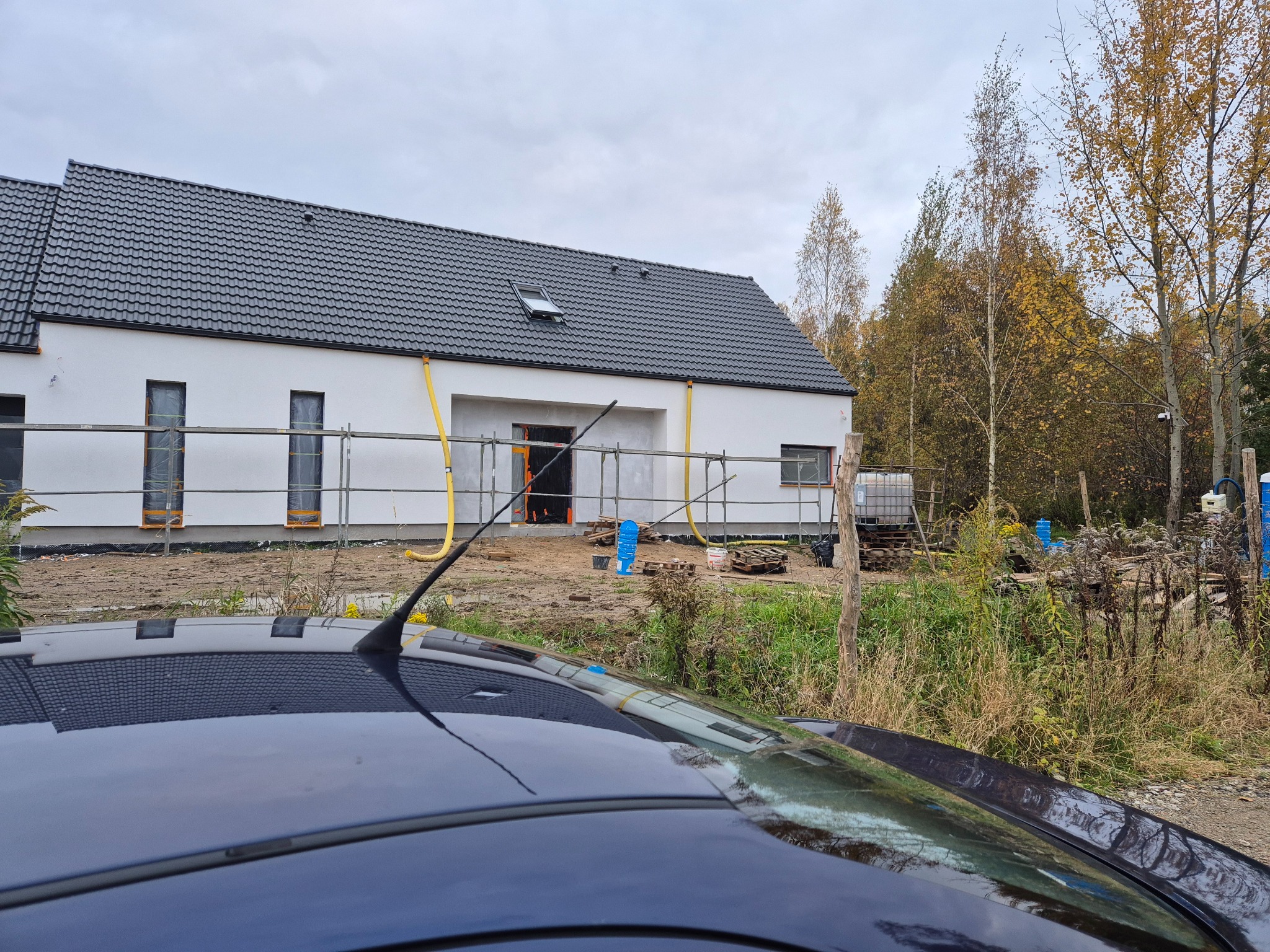 Biały dom z szarą dachówką w trakcie wykończenia, widok zza dachu samochodu. Widoczne rusztowania, okna zabezpieczone folią i elementy instalacji na elewacji.