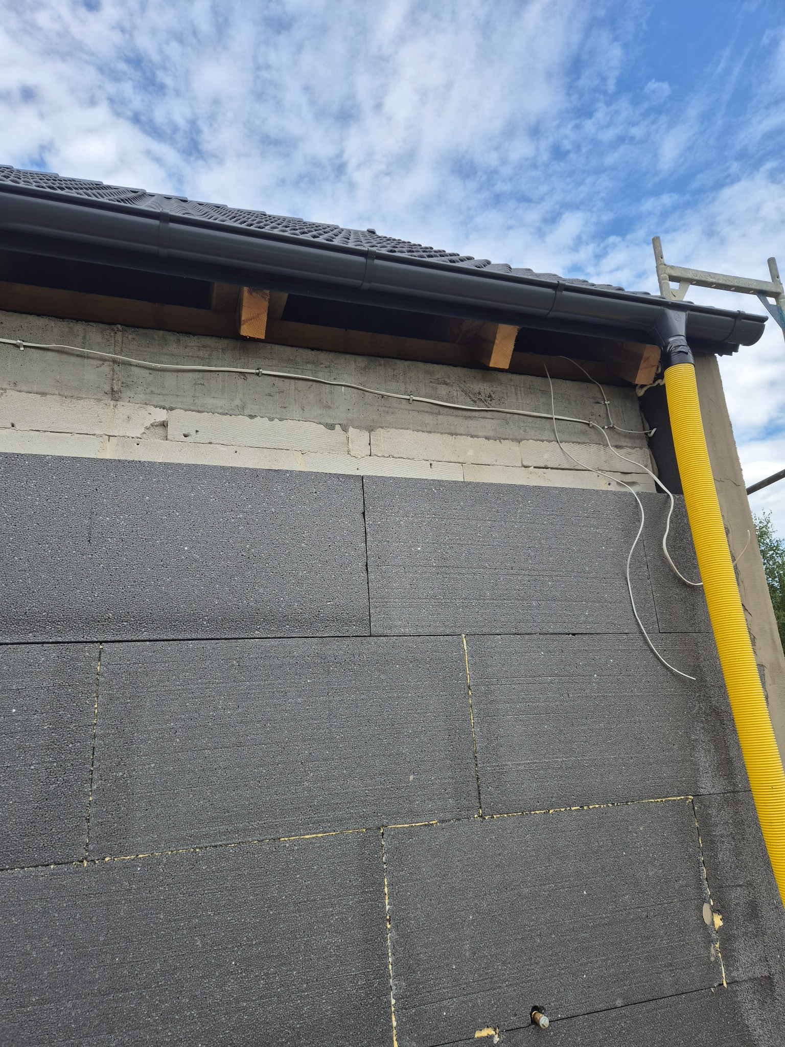 Ściana budynku w trakcie ocieplania: szare płyty styropianowe, widoczne mocowania, fragment dachu z rynną i żółta rura wentylacyjna na tle błękitnego nieba.