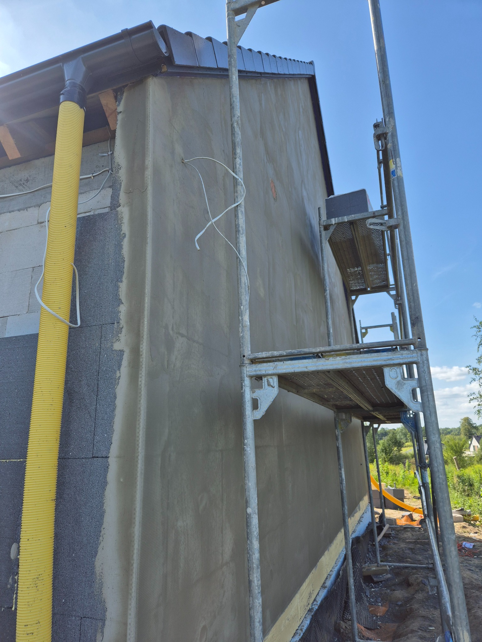 Ściana budynku w trakcie szpachlowania, widoczne rusztowanie i żółta rura wentylacyjna. Świeżo nałożona warstwa szpachli na siatce. Prace wykończeniowe na zewnątrz.