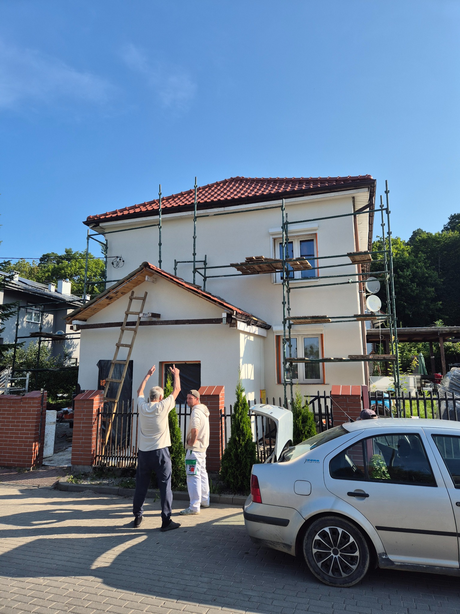 Dwóch mężczyzn przy rusztowaniu maluje elewację domu jednorodzinnego w słoneczny dzień. Widoczna drabina i samochód osobowy. Prace w toku.