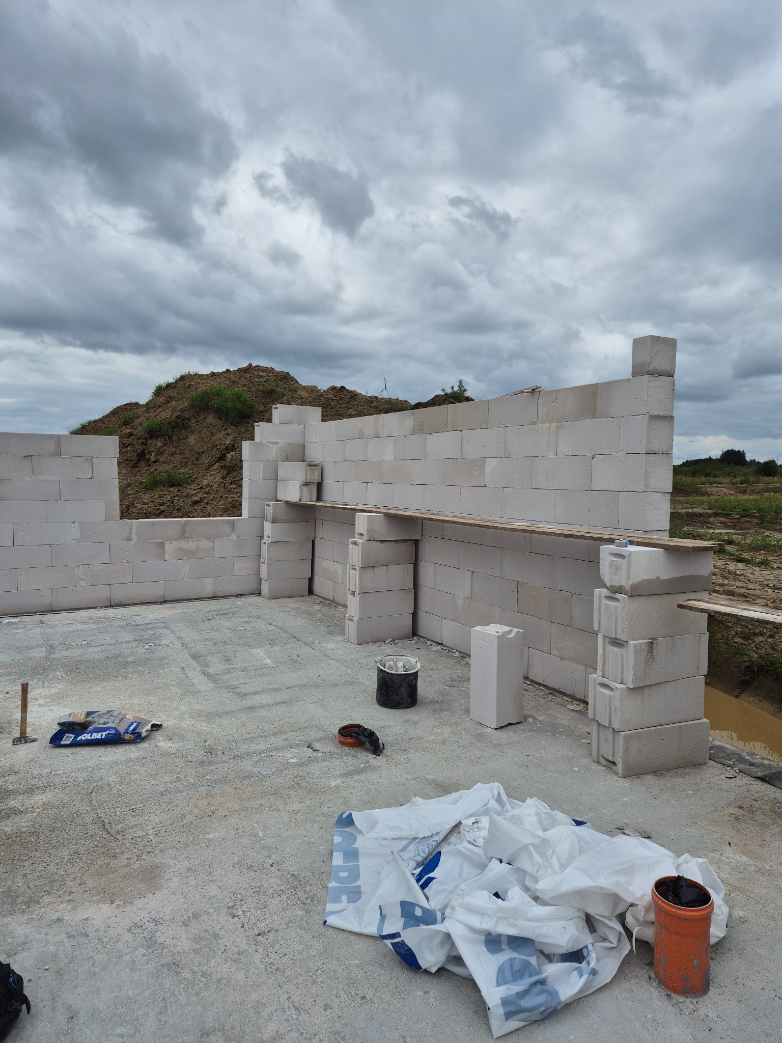 Wznoszenie ścian z bloczków betonu komórkowego na fundamencie. Widoczne narzędzia i materiały budowlane. Pochmurne niebo w tle. Etap budowy domu.