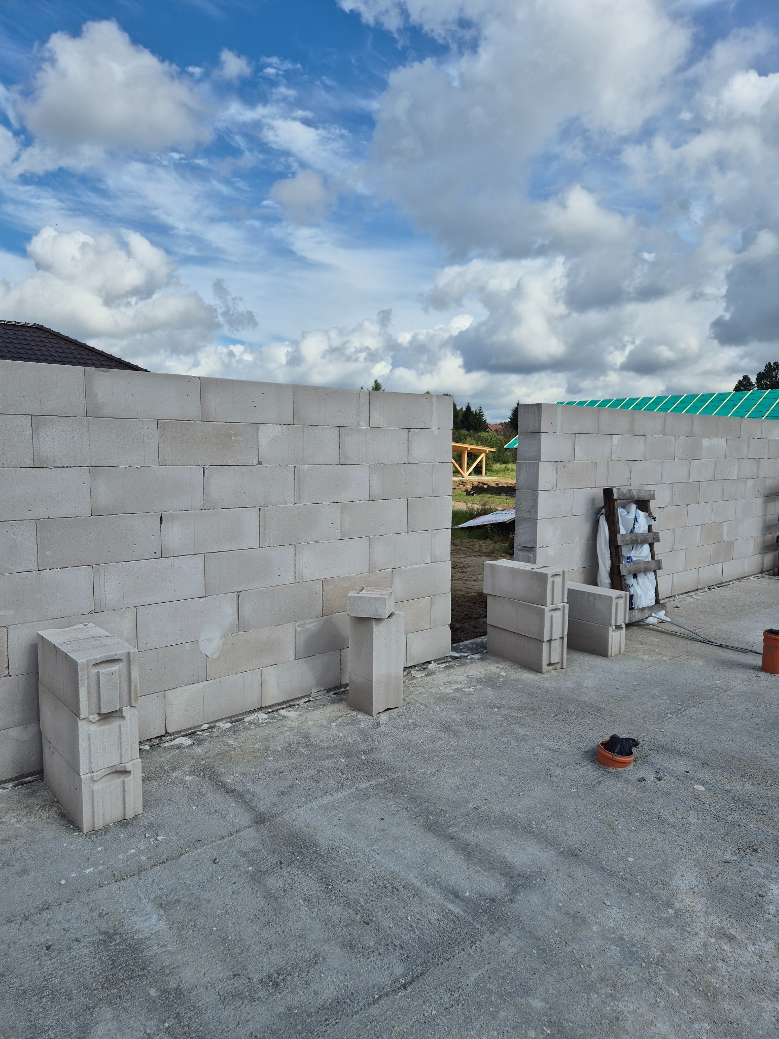 Wznoszenie ściany z betonu komórkowego Ytong na fundamencie. Widoczne bloczki, drabina oparta o ścianę i fragment dachu w tle. Budowa w toku, słoneczny dzień.