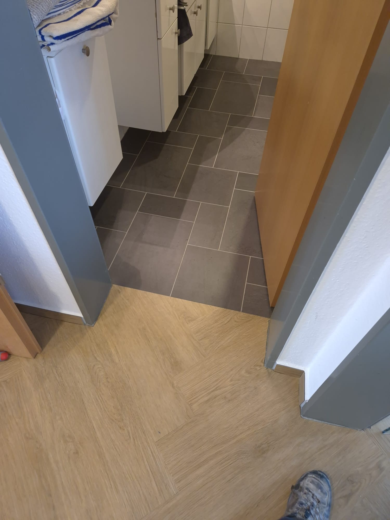 Połączenie dwóch rodzajów podłóg: parkiet w jodełkę i szare płytki w łazience. Widoczna dolna część futryny drzwi i fragment buta.