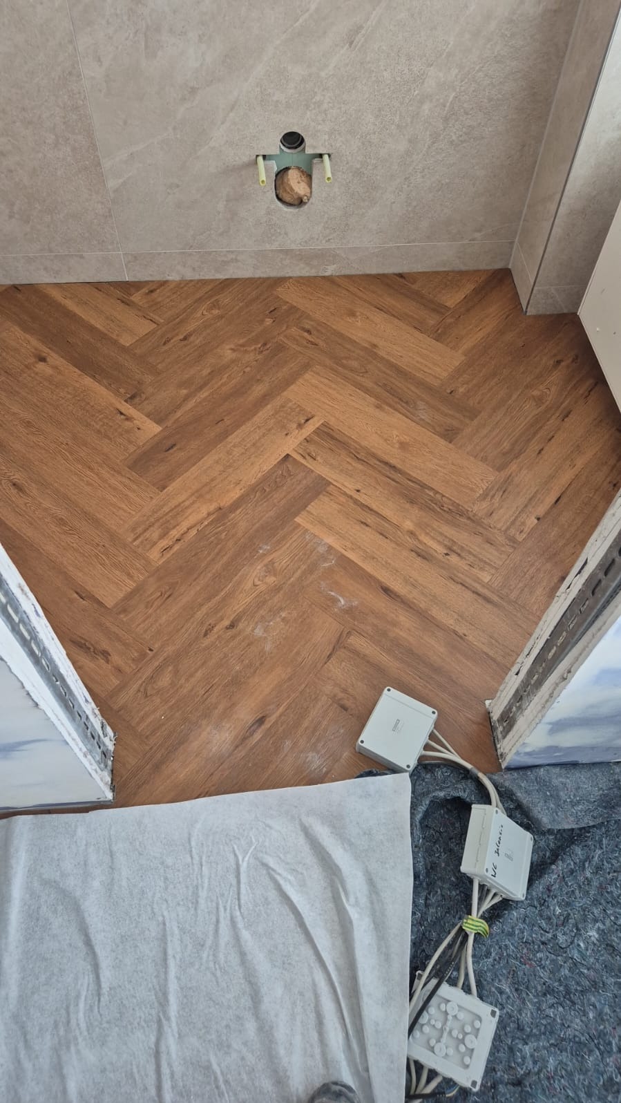 Ukończona podłoga z paneli ułożonych w jodełkę, widok z góry. Na ścianie szara płytka, widoczne puszki elektryczne i zabezpieczenia podłogi.