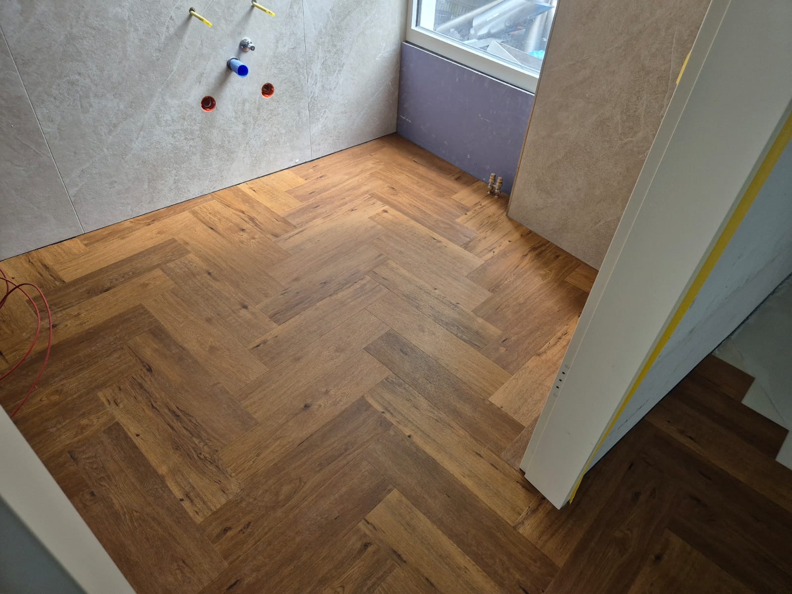 Ukończona podłoga z paneli ułożonych w jodełkę w łazience z częściowo wykończonymi ścianami. Widoczne przyłącza wodne i fragment okna.