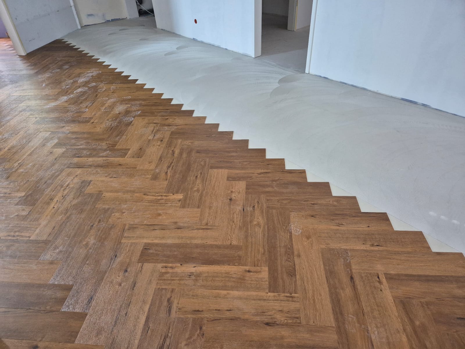 Ułożona podłoga z paneli w jodełkę, kontrastująca z przygotowanym podłożem pod dalsze prace. Widoczne białe ściany i fragmenty otworów drzwiowych.