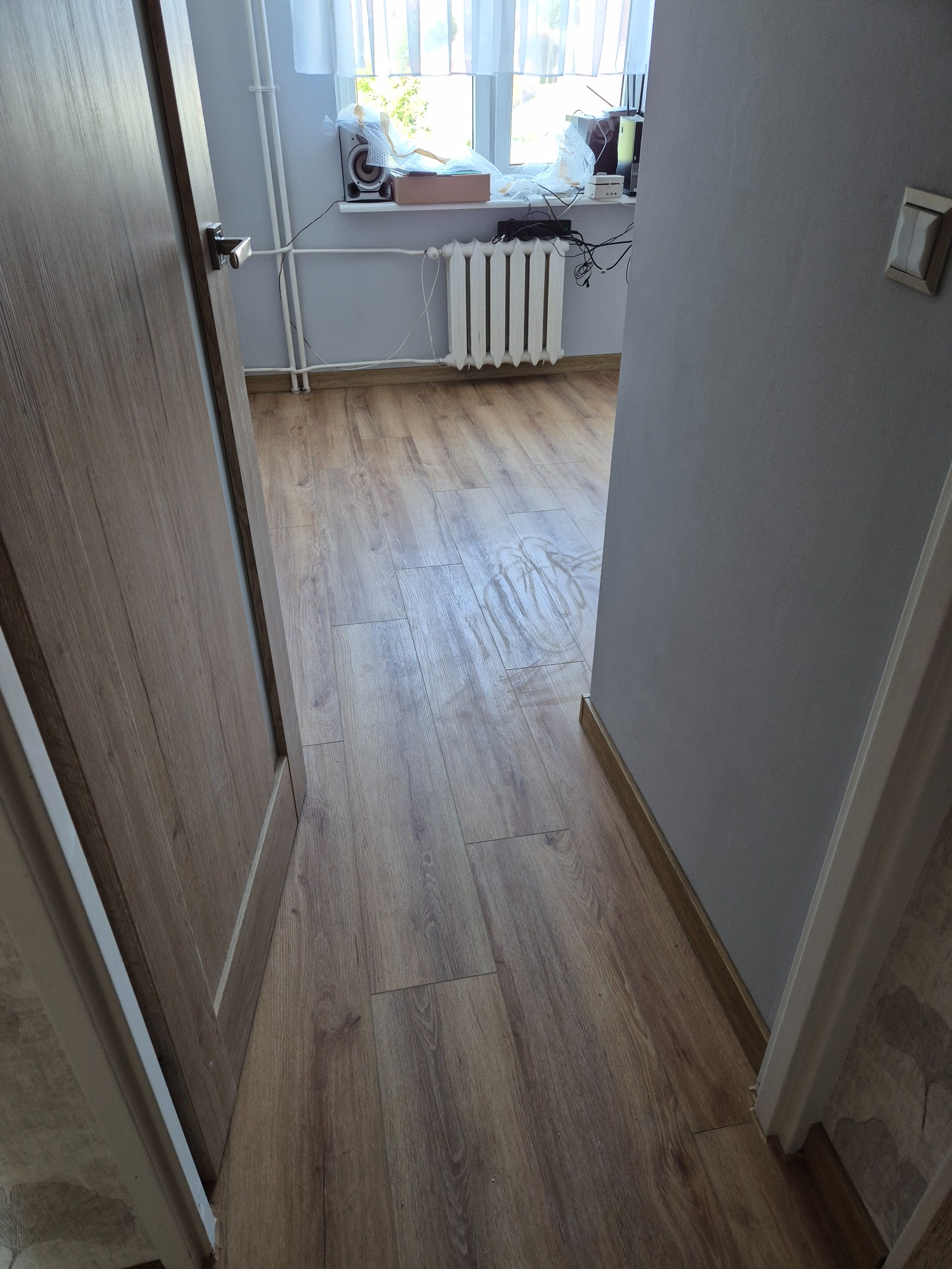 Widok zza uchylonych drzwi na odnowione wnętrze: drewniana podłoga, szare ściany, biały grzejnik pod oknem. Perspektywa zza framugi.