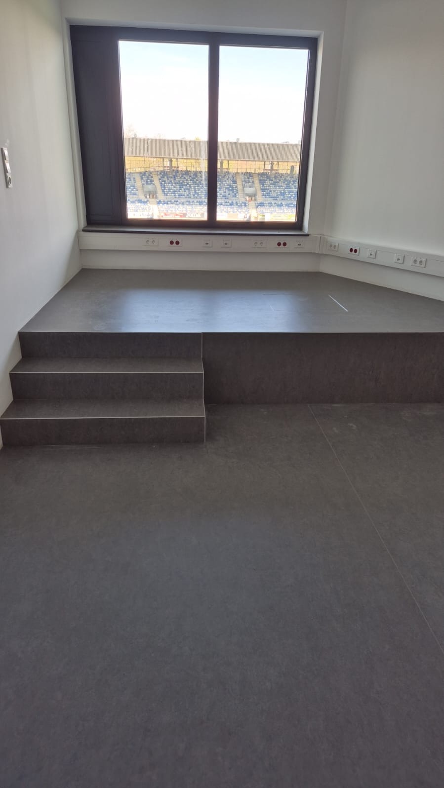 Szara, matowa podłoga z trzema schodkami prowadzącymi na podest. W tle okno z widokiem na stadion. Białe ściany i listwy przypodłogowe.