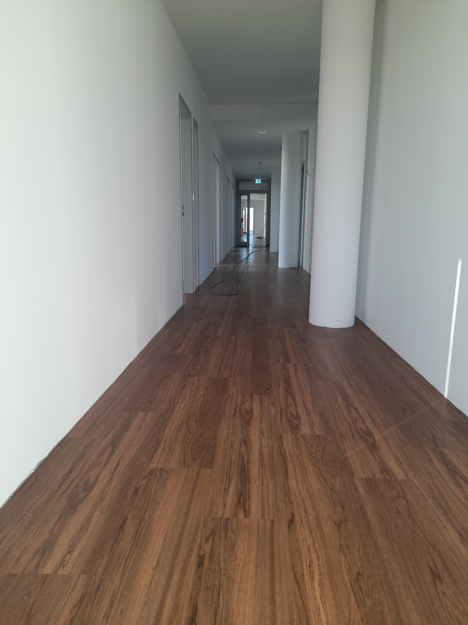 Długi korytarz z panelami podłogowymi w odcieniach brązu i białymi ścianami. Widoczne drzwi po lewej stronie i kolumny po prawej. Na końcu korytarza widoczne światło.