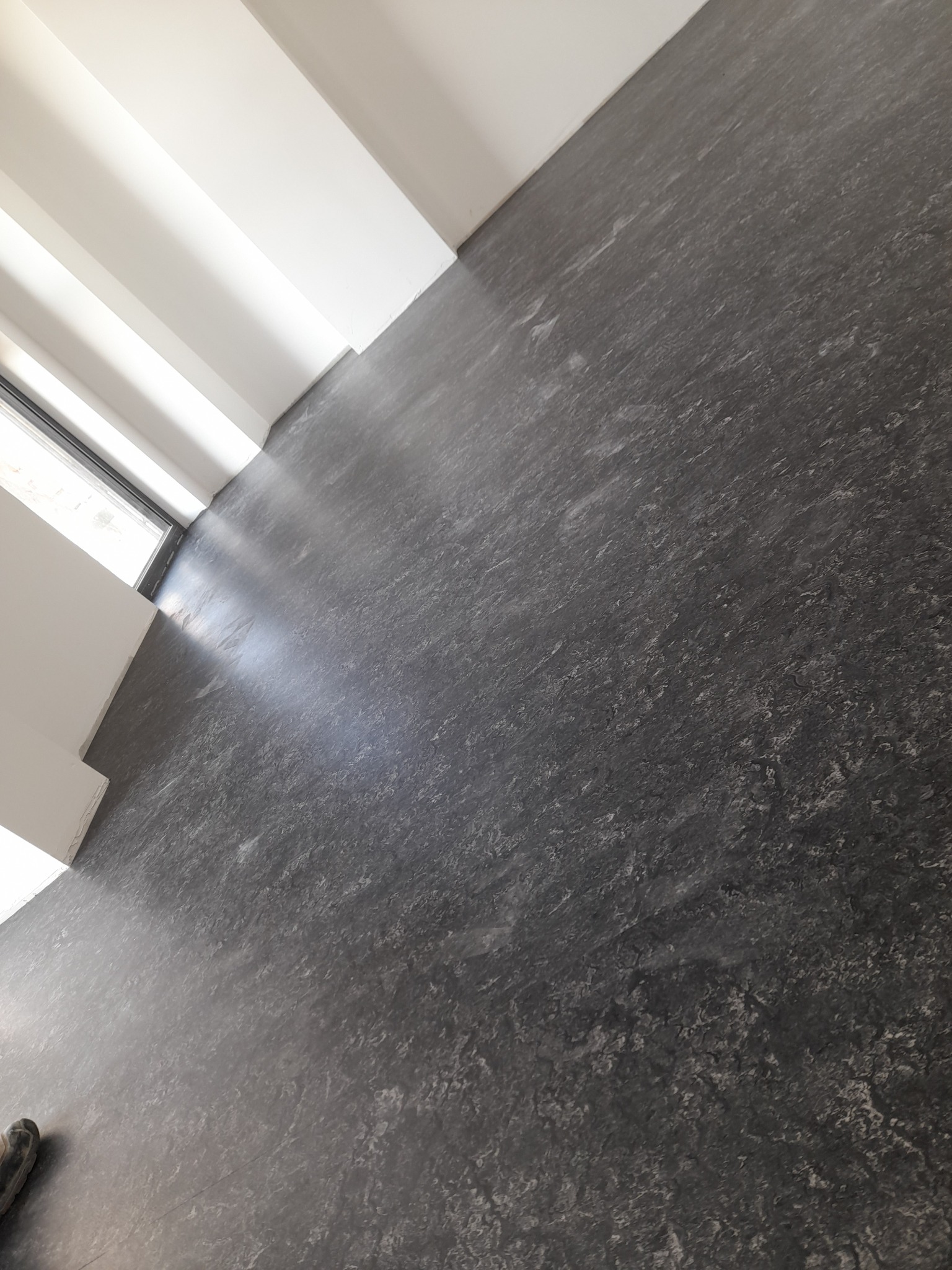 Szara, wzorzysta podłoga w jasnym pomieszczeniu z oknem. Widoczny fragment buta w lewym dolnym rogu. Ujęcie pod kątem, podkreślające fakturę podłogi.