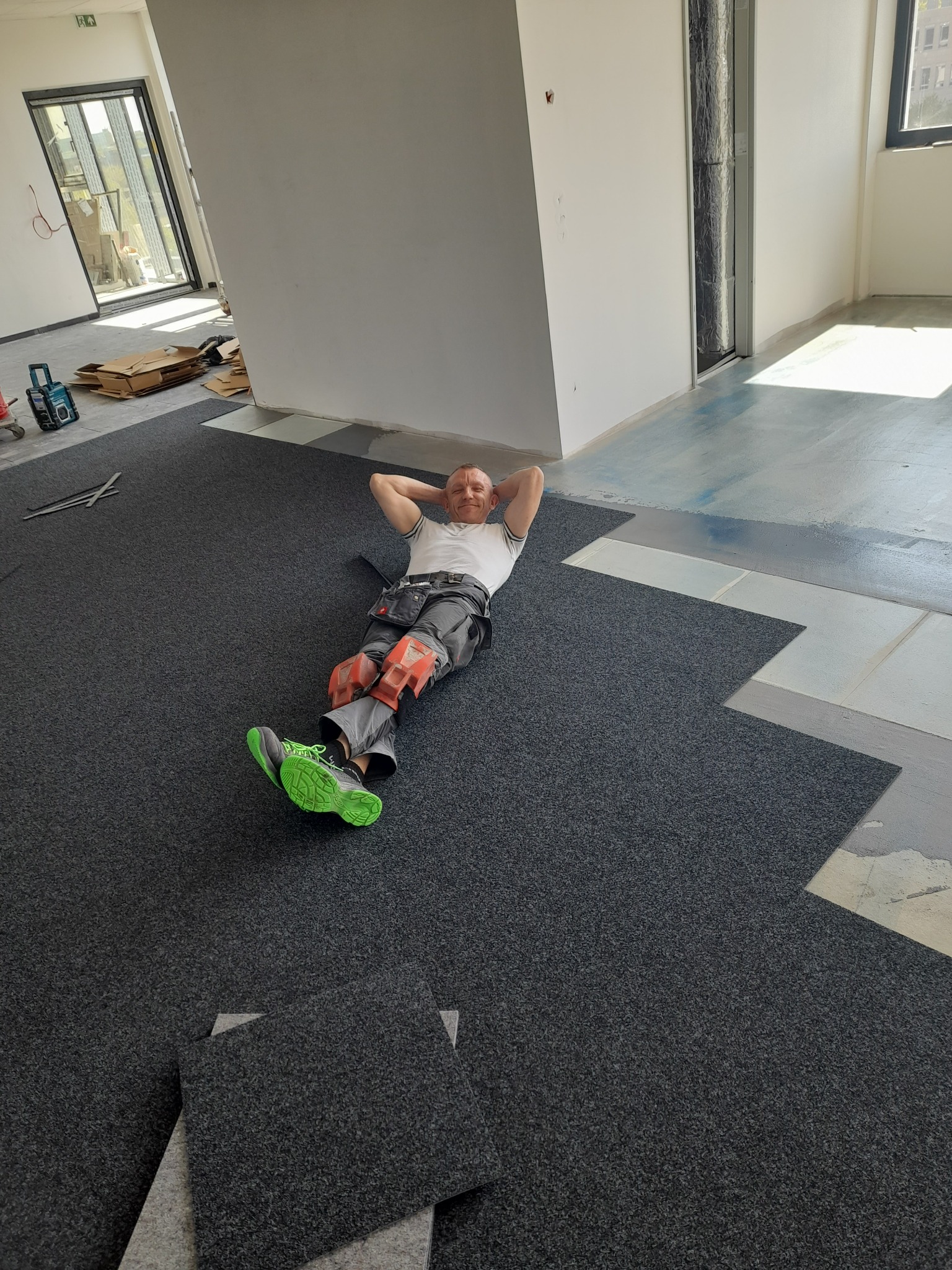 Układanie ciemnych paneli podłogowych w jasnym wnętrzu. Na pierwszym planie uśmiechnięty mężczyzna w roboczym stroju, odpoczywający na ułożonej podłodze. Widoczne narzędzia.