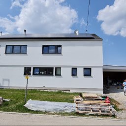 ELISA BAU SPÓŁKA Z OGRANICZONĄ ODPOWIEDZIALNOŚCIĄ - Biały, dwukondygnacyjny dom z ciemnymi oknami po malowaniu elewacji. Na dachu panele słoneczne. Na pierwszym planie gruz i materiały budowlane, a w tle błękitne niebo.