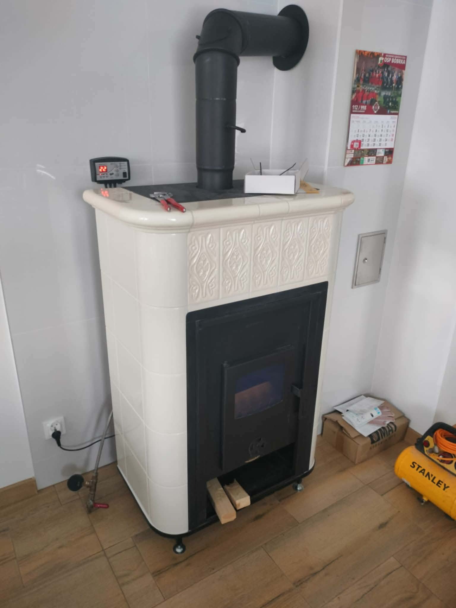 Biały piec kaflowy z czarnym wkładem kominkowym i rurą, widoczny regulator temperatury, narzędzia na górze, podłoga drewniana, ściany białe, kalendarz na ścianie.