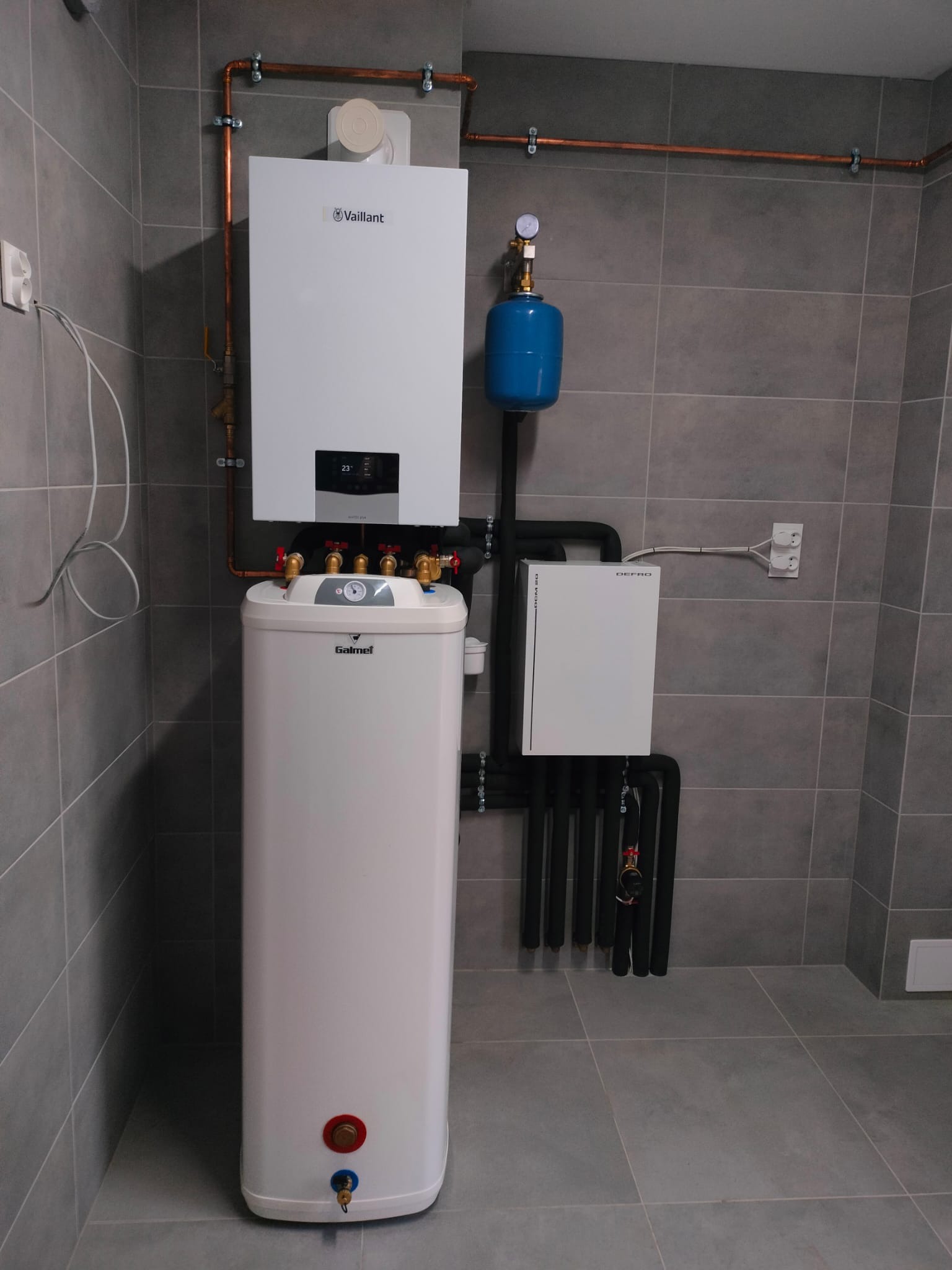 Biały piec gazowy Vaillant i zasobnik Galmet na tle szarych płytek. Miedziane rury i czarne przewody tworzą instalację. Widoczny manometr i skrzynka Defro.