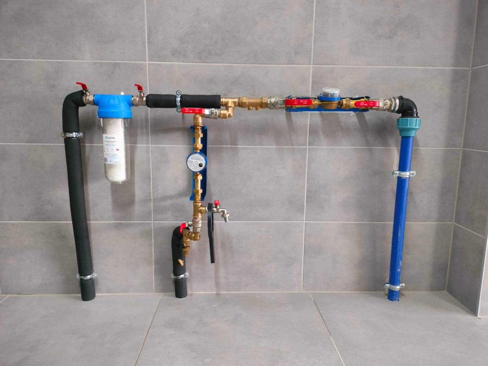 Kompletna instalacja wodna z filtrem, licznikiem i zaworami, zamontowana na szarej ścianie z płytek. Widoczne rury czarne, niebieskie i mosiężne.