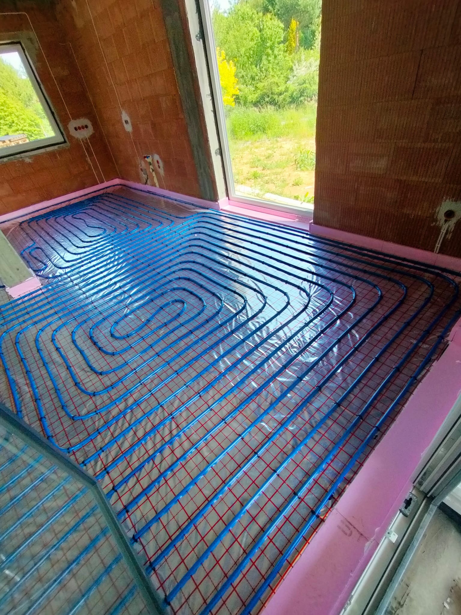 Niebieskie rury ogrzewania podłogowego ułożone na folii z czerwoną siatką, widok z góry w surowym wnętrzu z oknem na zieleń. Rury ułożone spiralnie.