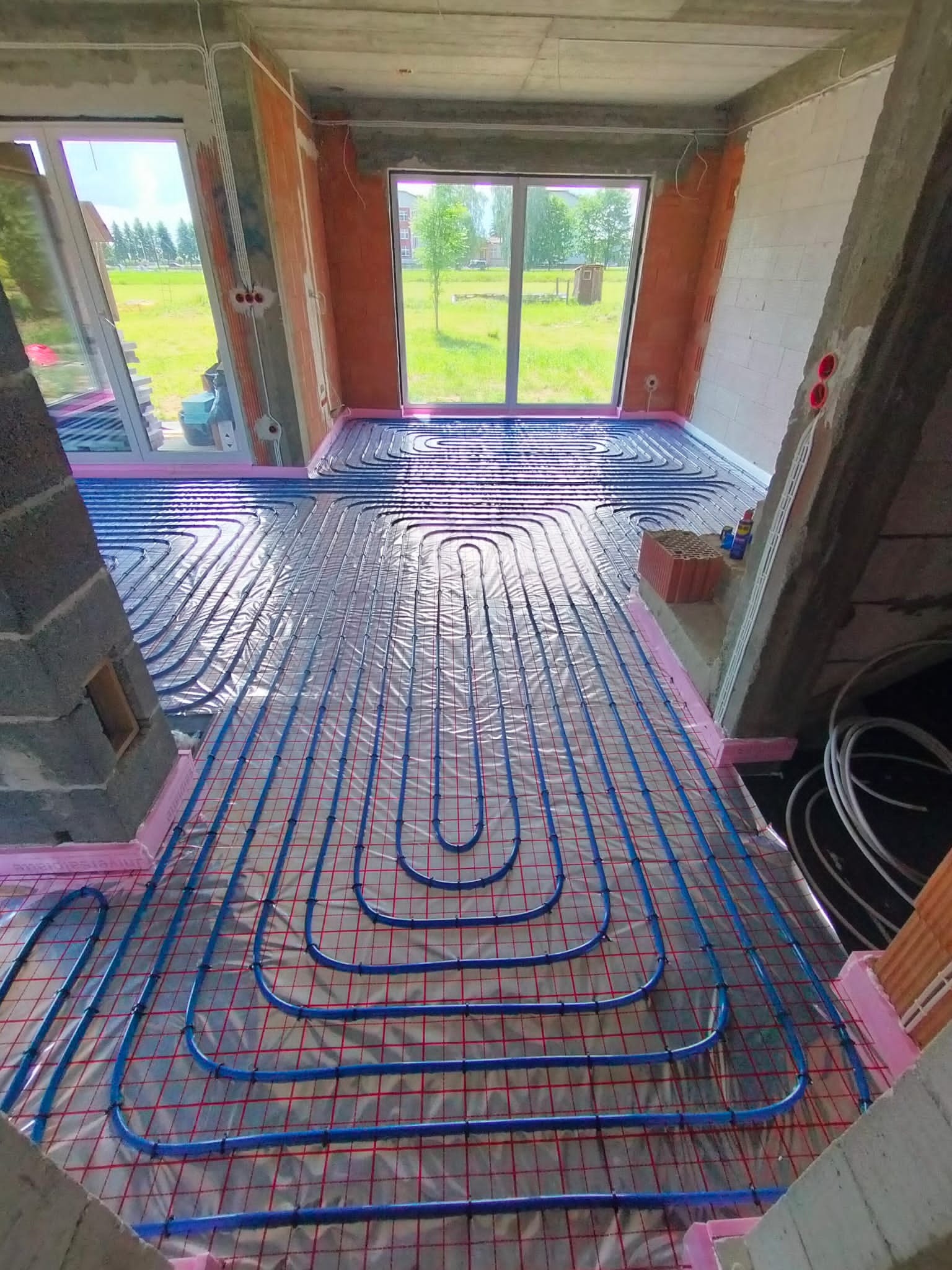 Niebieskie rury ogrzewania podłogowego ułożone na czerwonej siatce, widoczne przezroczyste folie, różowa izolacja przy ścianach, surowy stan budynku, okna z widokiem na zieleń.