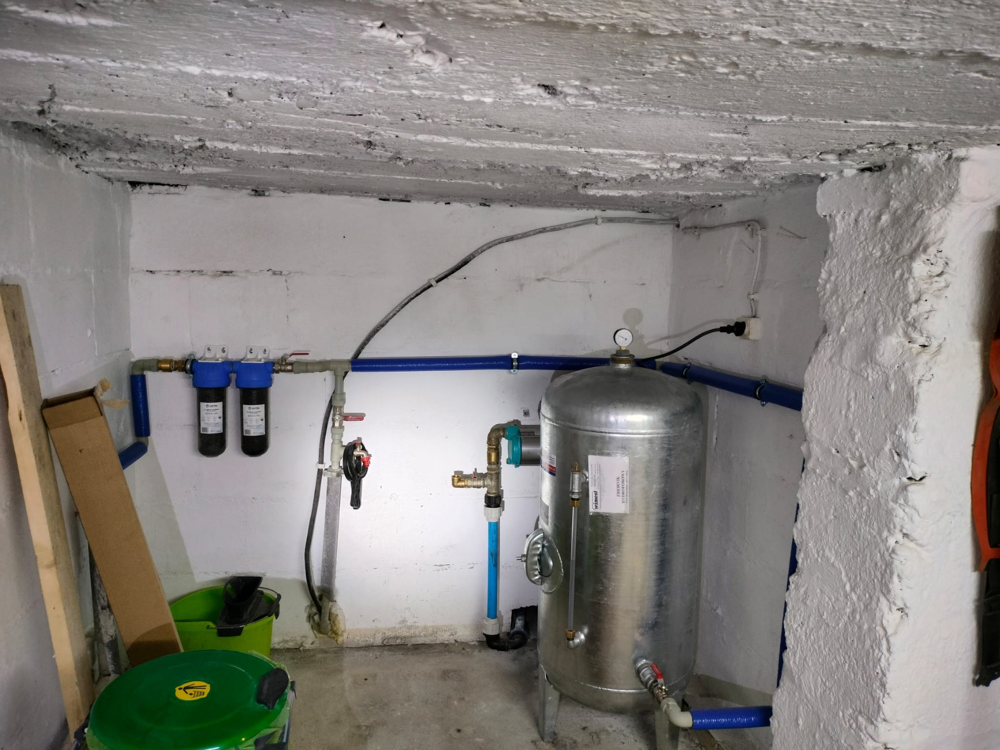 Hydrofor w piwnicy z filtrami wody i niebieskimi rurami. Widoczne narzędzia i akcesoria hydrauliczne. Ściany i sufit z surowym wykończeniem.