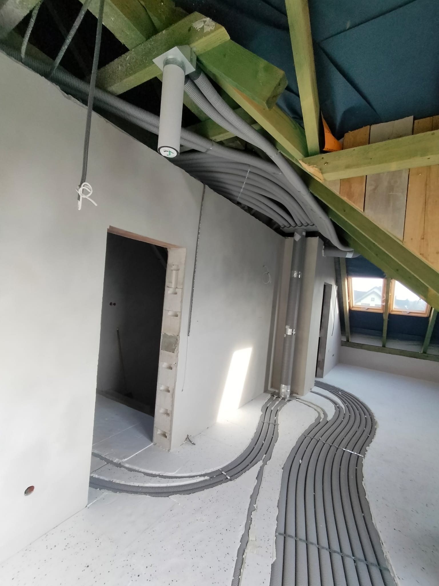 Szare, karbowane rury instalacyjne ułożone w bruzdach w podłodze na poddaszu, widoczne elementy konstrukcji dachu i fragment ściany z otworem drzwiowym.