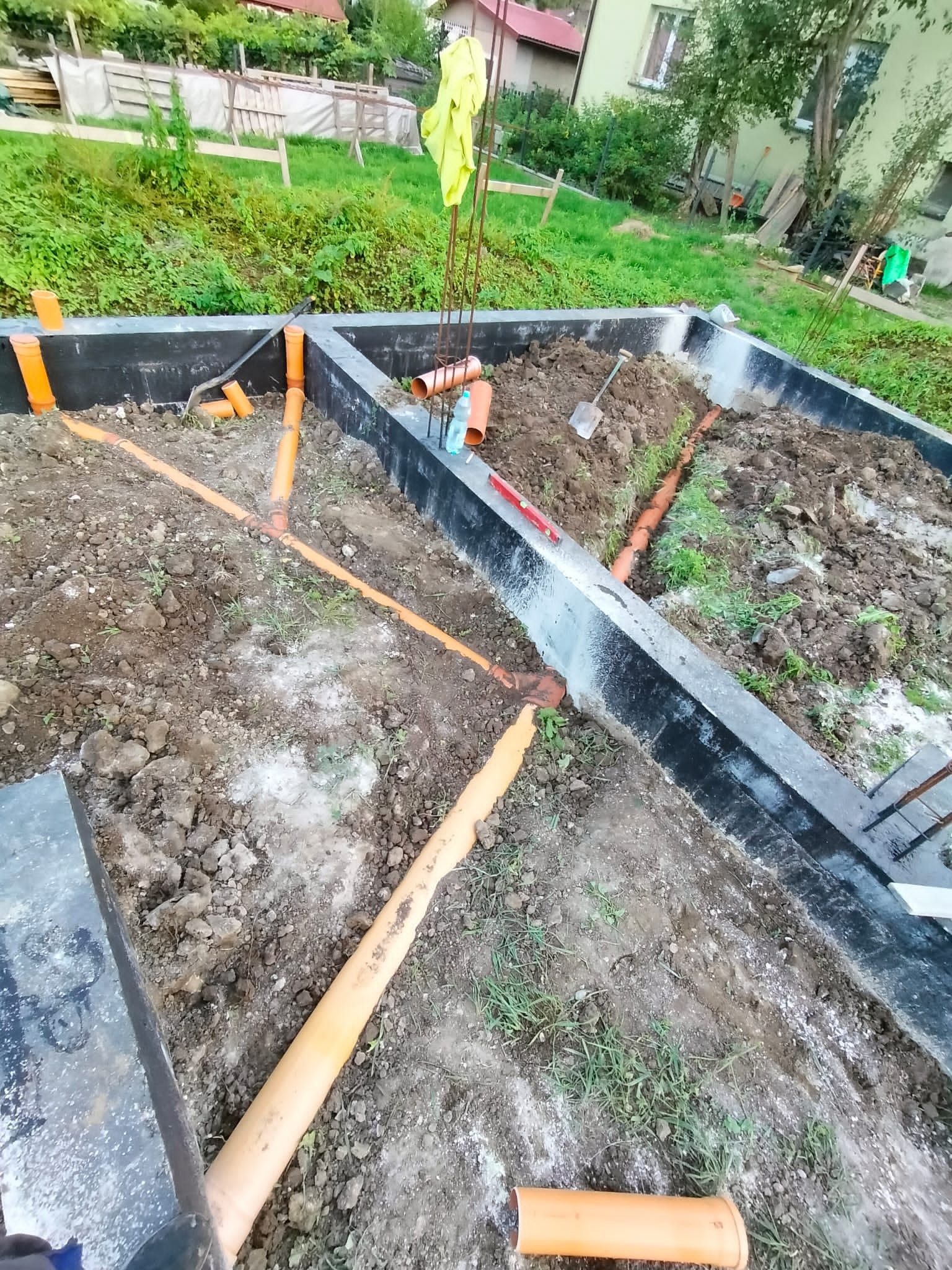Fundament budynku z ułożonymi pomarańczowymi rurami kanalizacyjnymi w ziemi. Widoczna łopata i poziomica. Tło stanowi zieleń i budynek.