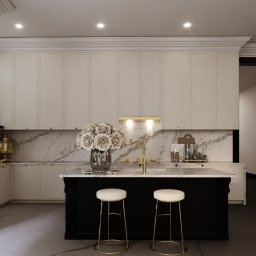 DNP Trading&Development Sp. z o.o. - Elegancka kuchnia z wyspą, białymi szafkami i marmurowym blatem. Na wyspie wazon z białymi różami i dwa złote hokery. Oświetlenie punktowe.