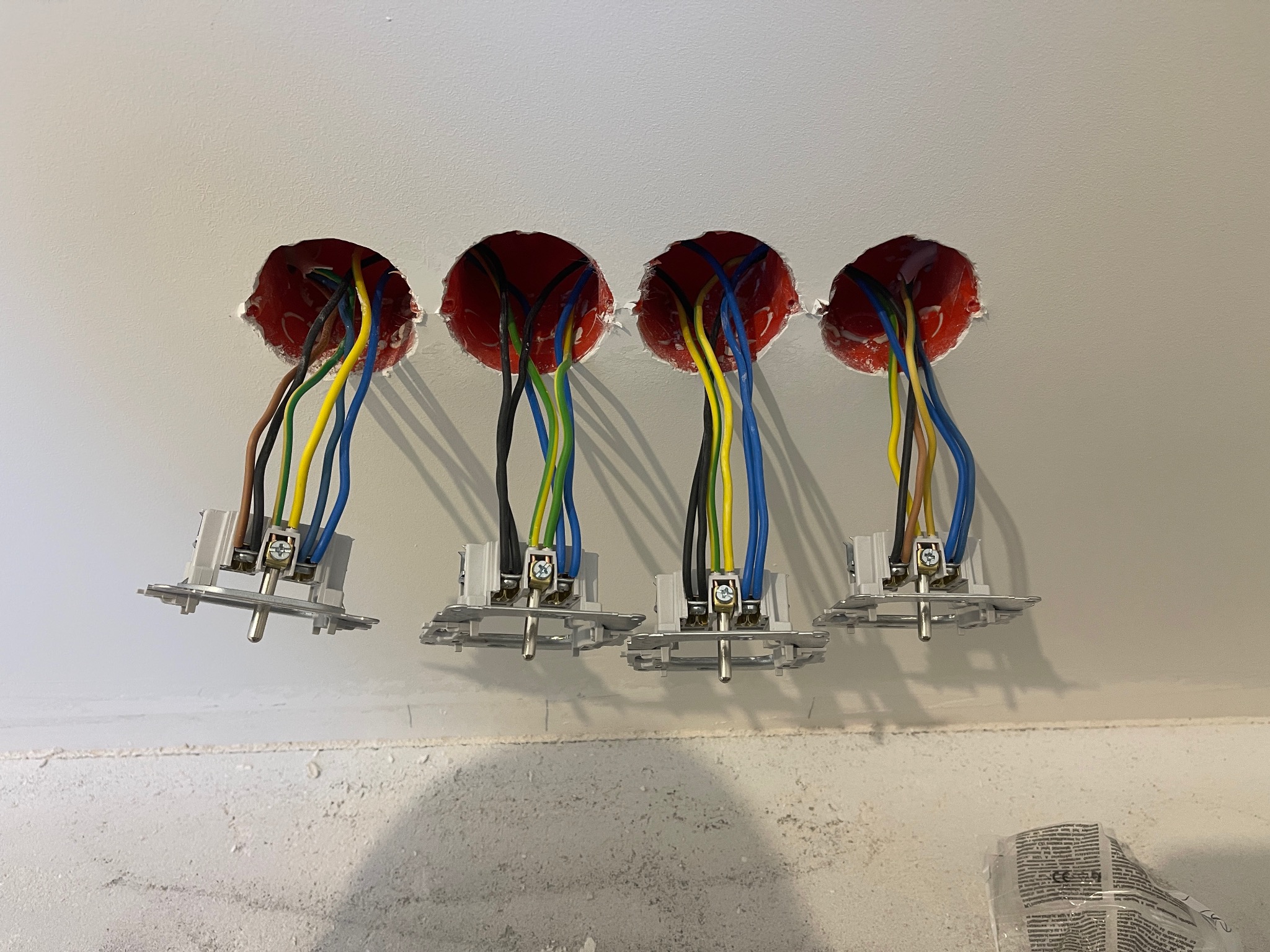 Cztery puszki instalacyjne z przewodami (brązowy, czarny, zielono-żółty, niebieski) i zamontowanymi gniazdkami, gotowe do osadzenia w ścianie. Widok z bliska.