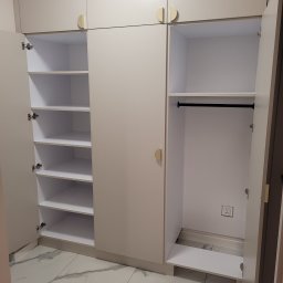 Jacek Stefańczyk NAS-TIME - Jasna szafa wnękowa z otwartymi drzwiami, ukazująca półki i drążek na ubrania. Widoczna marmurowa podłoga i gniazdko elektryczne wewnątrz szafy. Minimalistyczne, półokrągłe uchwyty.