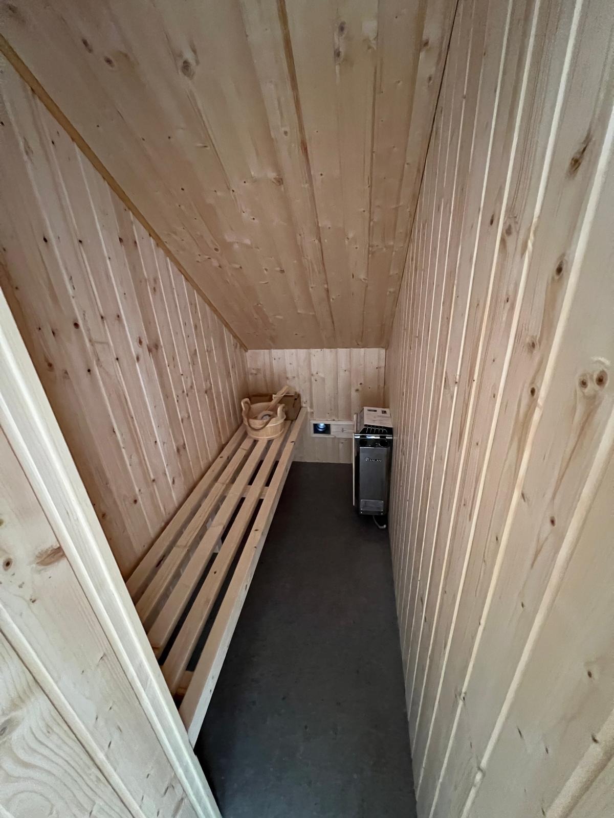 Wnętrze sauny z boazerią, drewnianymi ławkami i piecem. Ukośny sufit i oświetlenie tworzą przytulny klimat. Widoczny kubeł i chochla.