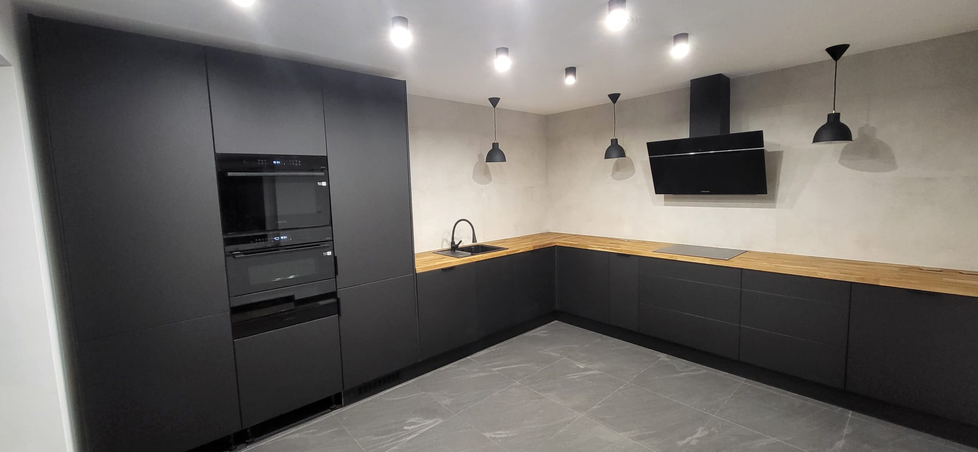Nowoczesna, minimalistyczna kuchnia z czarnymi matowymi frontami i drewnianym blatem. Uwagę przykuwają wiszące lampy i sprzęt AGD w zabudowie.