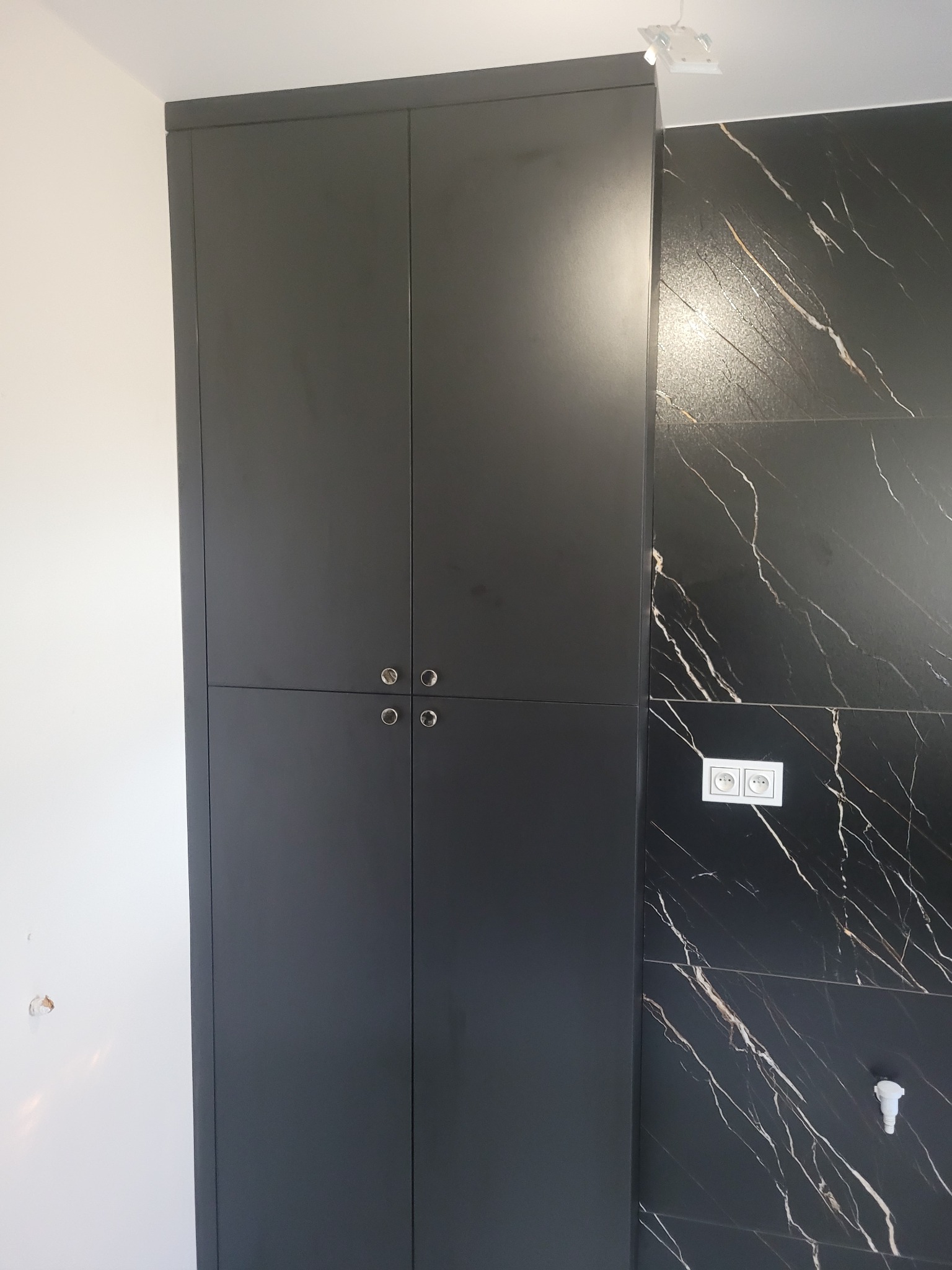 Czarna, wysoka szafa wnękowa z czterema drzwiami i okrągłymi uchwytami, na tle ściany z czarnych płytek imitujących marmur. Widoczne gniazdka elektryczne.