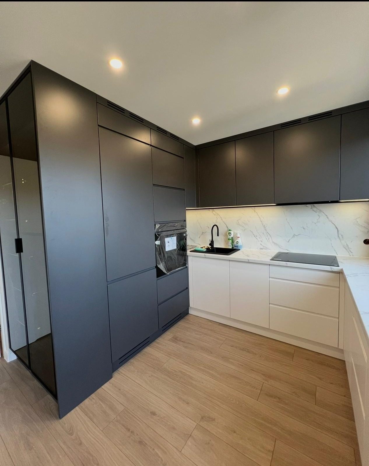 Nowoczesna kuchnia z szarymi szafkami górnymi i białymi dolnymi, marmurowym blatem i czarną armaturą. Widoczny piekarnik w zabudowie i płyta indukcyjna.