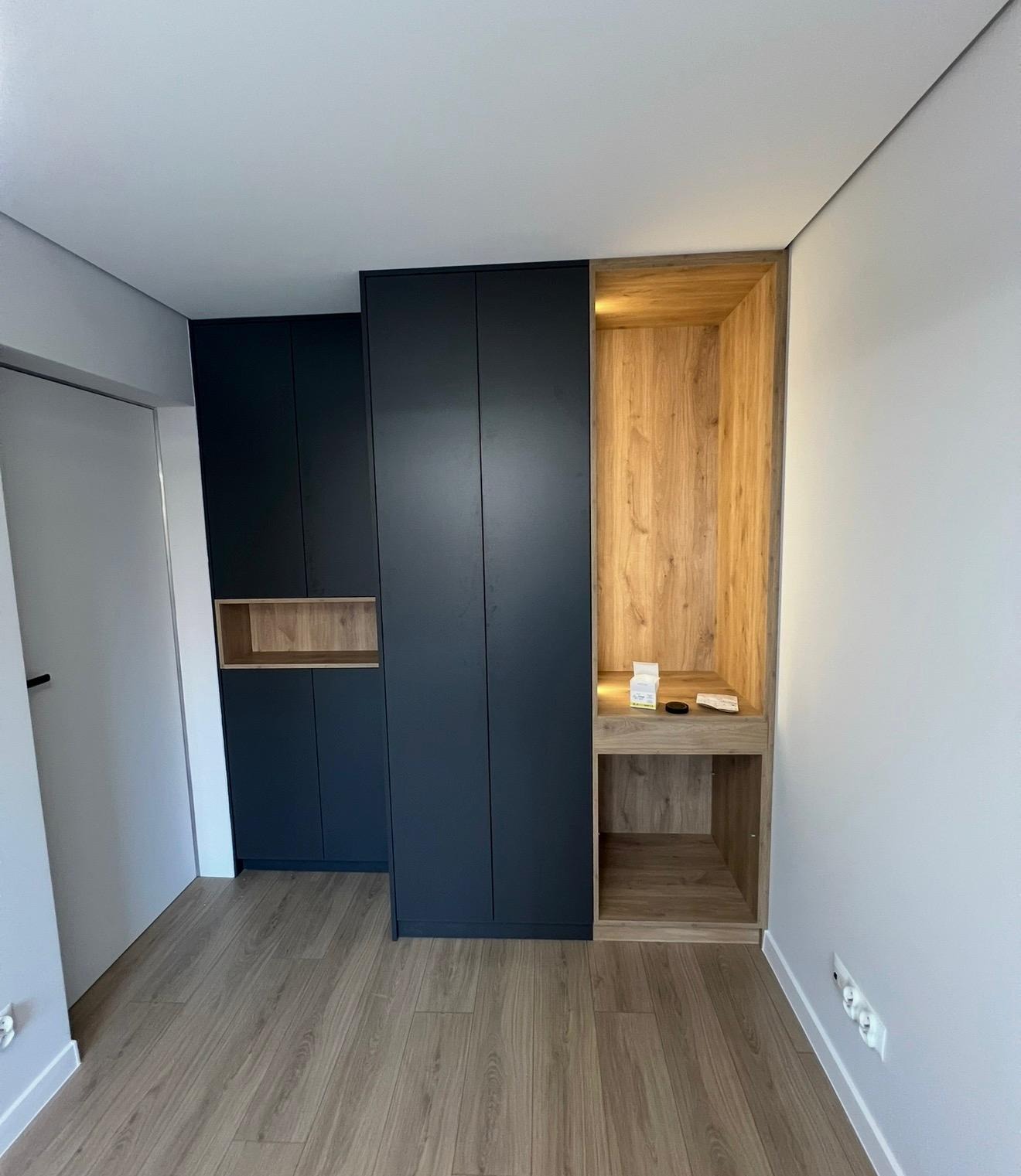 Nowoczesna szafa wnękowa z ciemnymi frontami i drewnianą wnęką z półkami, dopasowana do jasnego wnętrza z drewnianą podłogą. Minimalistyczny design.