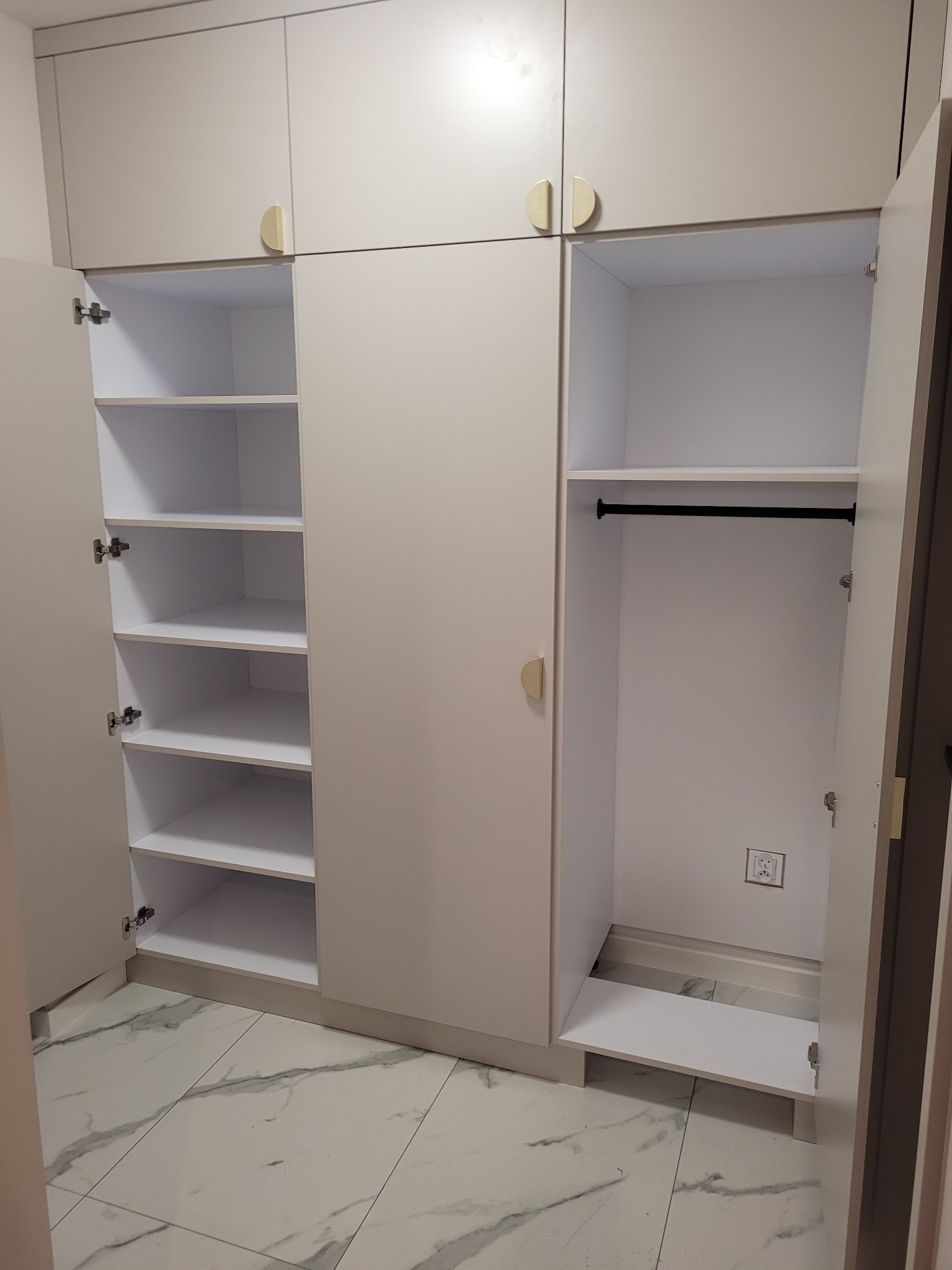Jasna szafa wnękowa z otwartymi drzwiami, ukazująca półki i drążek na ubrania. Widoczna marmurowa podłoga i gniazdko elektryczne wewnątrz szafy. Minimalistyczne, półokrągłe uchwyty.