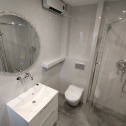 Marcin Herbut - Wykończona łazienka w szarościach: umywalka z szafką, toaleta podtynkowa, kabina prysznicowa z chromowanymi detalami i okrągłe lustro z kryształkami.