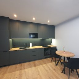 Marcin Herbut - Nowoczesna kuchnia w odcieniach szarości z drewnianym blatem i stołem. Minimalistyczny design, oświetlenie LED pod szafkami, drewniana podłoga.