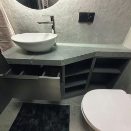 Marcin Herbut - Nowoczesna łazienka z umywalką nablatową, szarą zabudową meblową z otwartymi półkami i czarnym dywanikiem. Minimalistyczny design, płytki imitujące kamień.