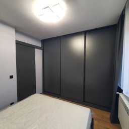 Marcin Herbut - Nowoczesna szafa przesuwna w ciemnym kolorze, idealnie wkomponowana w jasne wnętrze sypialni. Minimalistyczny design, gładkie fronty, subtelne oświetlenie sufitowe.