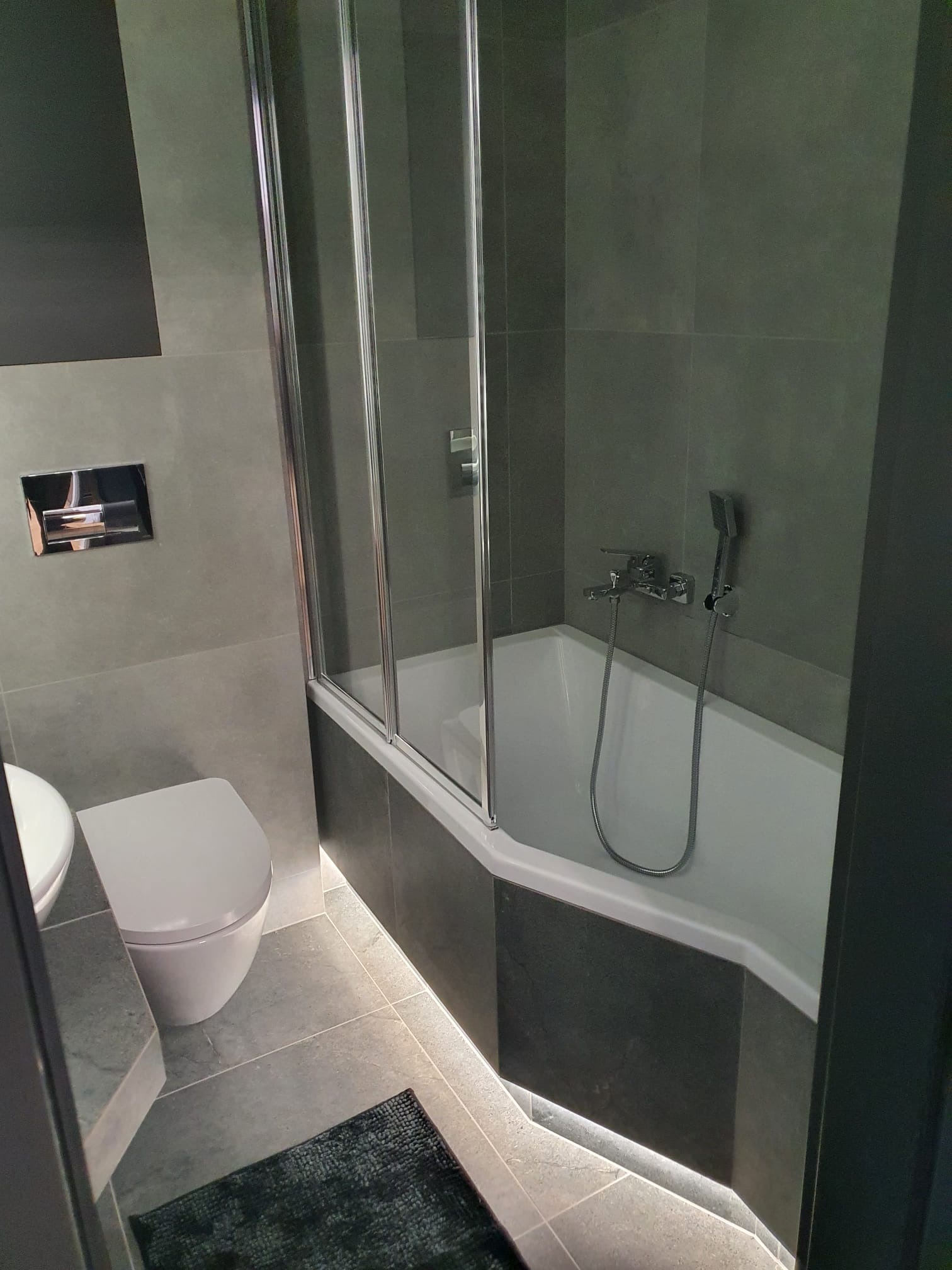 Nowoczesna łazienka w szarościach z podświetlaną wanną i kabiną prysznicową. Uwagę przykuwa minimalistyczna toaleta i ciemny dywanik.