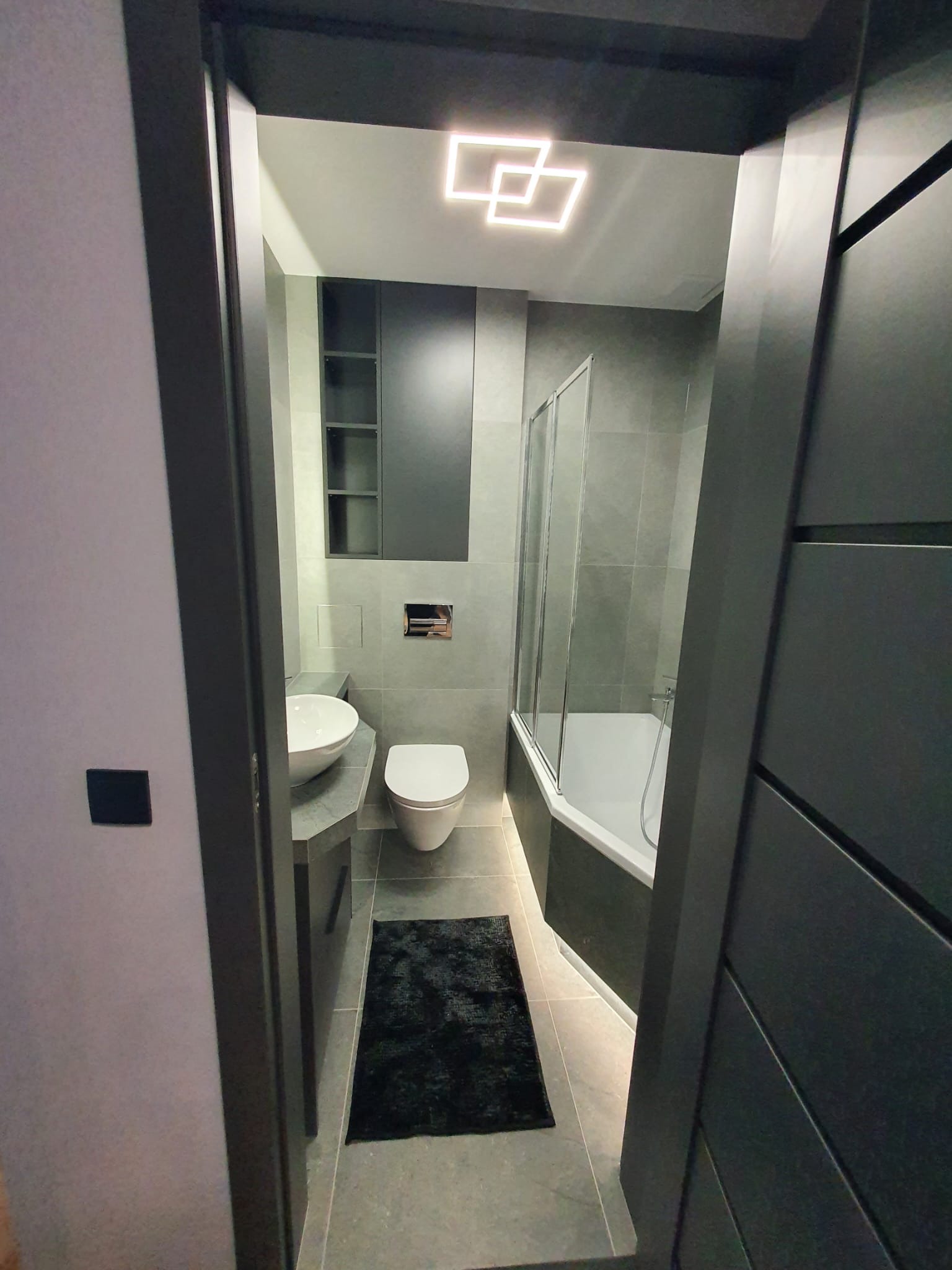 Nowoczesna łazienka w szarościach z geometrycznym oświetleniem sufitowym, kabiną prysznicową i umywalką na blacie. Widok zza futryny drzwi.