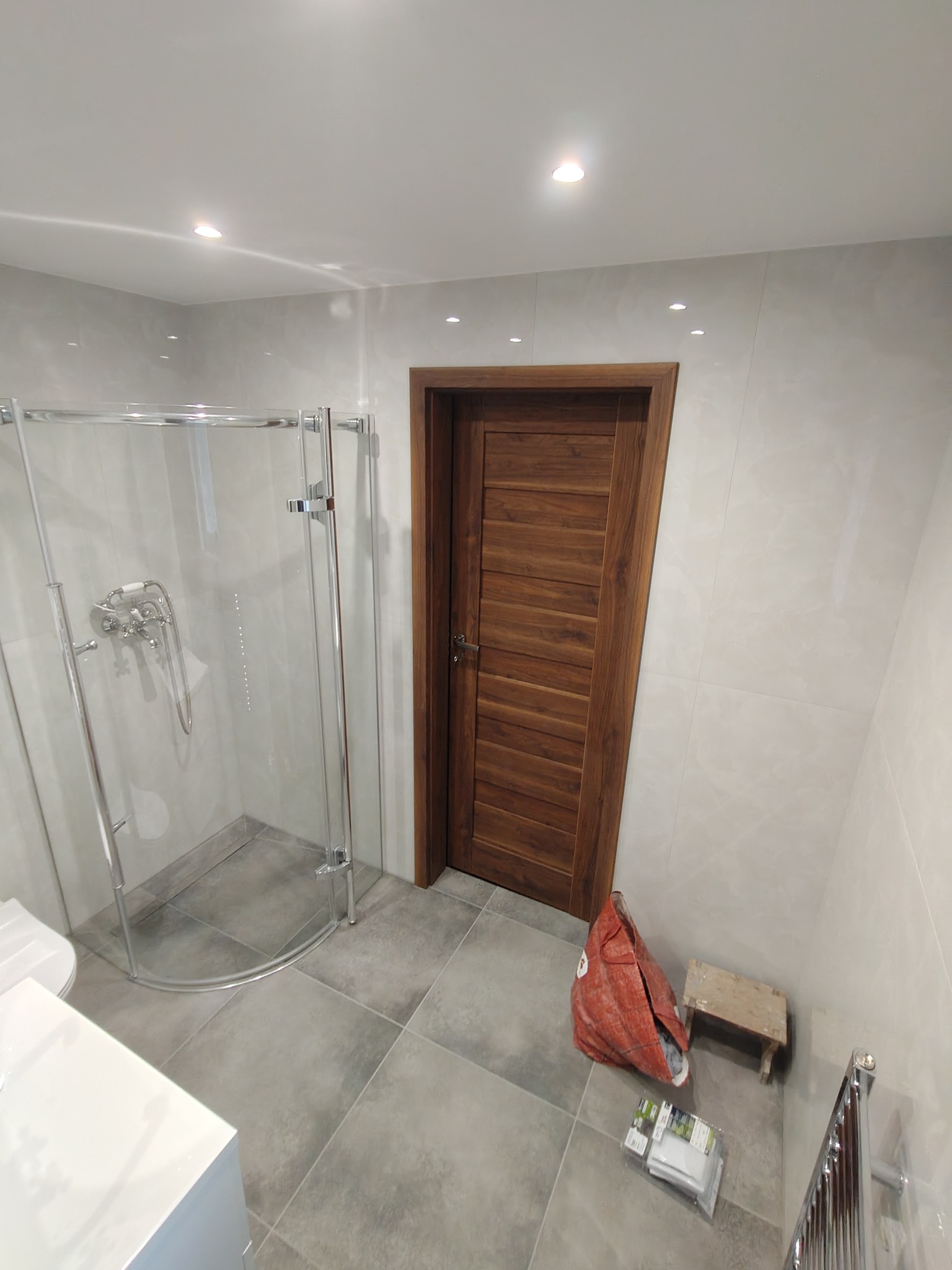 Wykończona łazienka z kabiną prysznicową, drewnianymi drzwiami i szarą glazurą. Widoczny worek budowlany i stołek, sugerujące prace wykończeniowe.