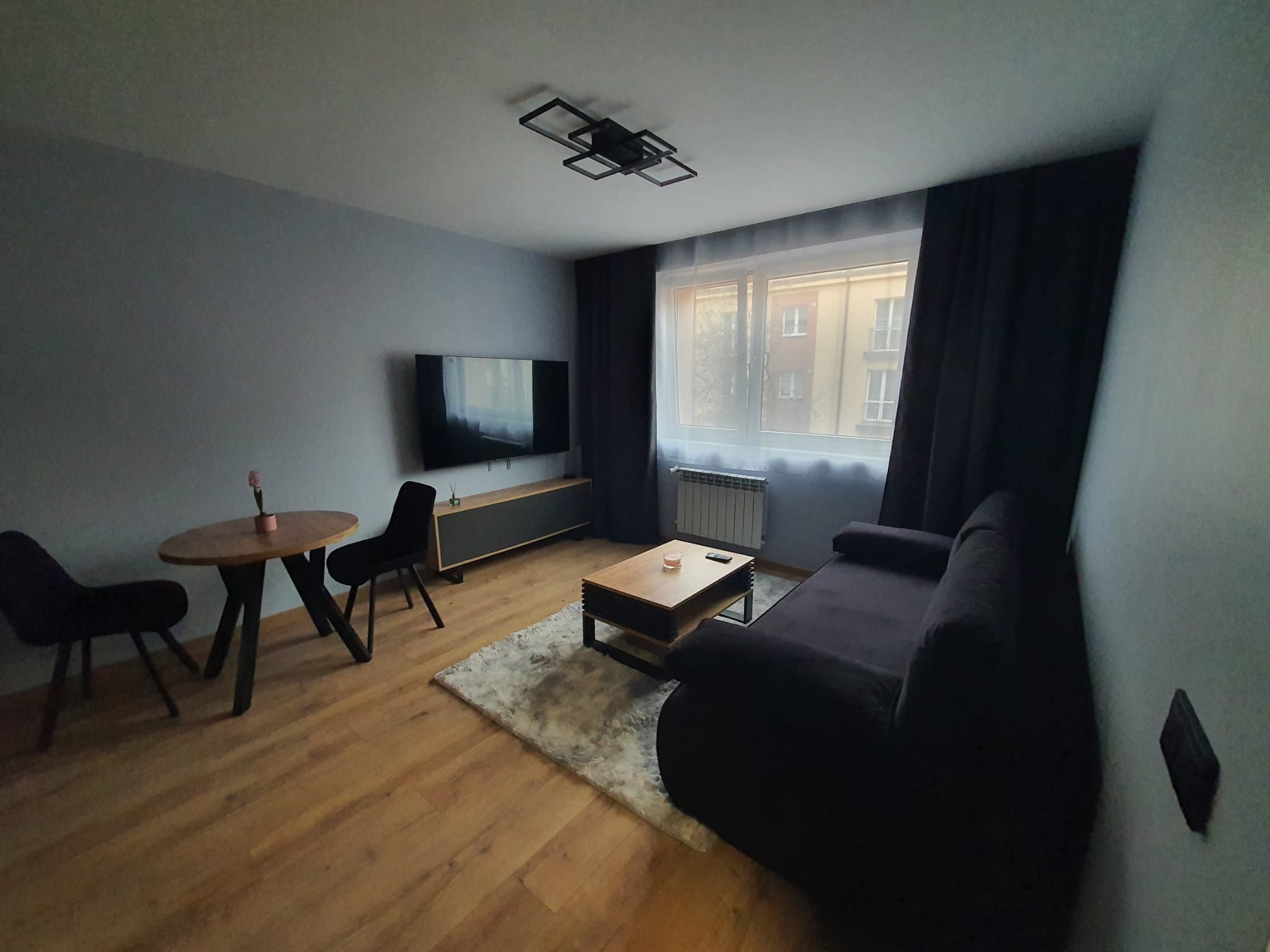 Nowoczesny salon po wykończeniu: drewniana podłoga, szara ściana, czarna sofa i krzesła, okrągły stół z kwiatem, dywan shaggy i designerska lampa sufitowa.