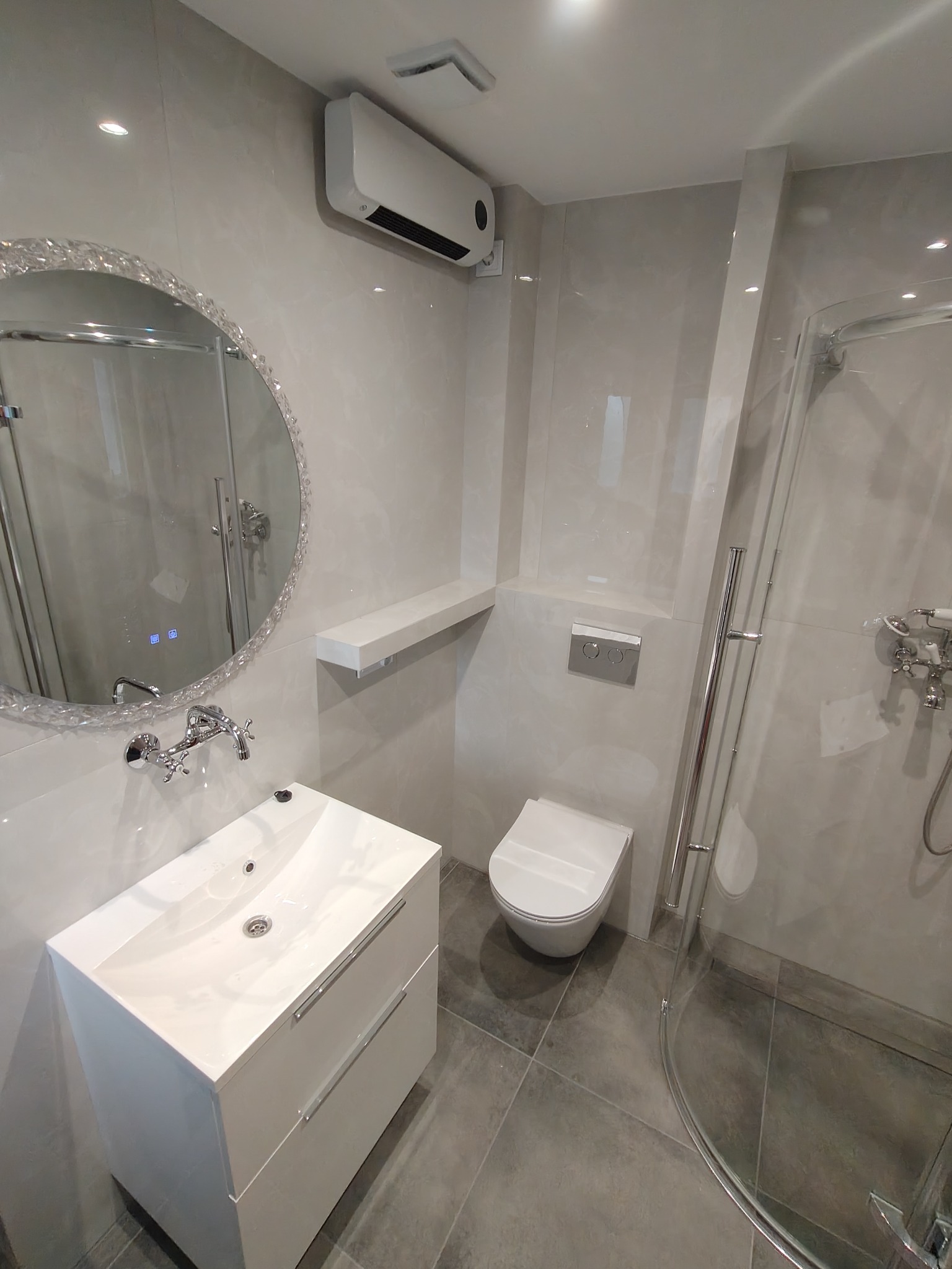 Wykończona łazienka w szarościach: umywalka z szafką, toaleta podtynkowa, kabina prysznicowa z chromowanymi detalami i okrągłe lustro z kryształkami.