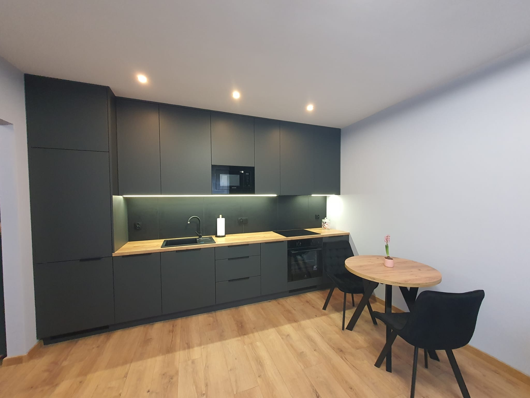 Nowoczesna kuchnia w odcieniach szarości z drewnianym blatem i stołem. Minimalistyczny design, oświetlenie LED pod szafkami, drewniana podłoga.